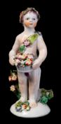 A Bow figure of a putto: holding a basket of flowers and wearing a floral headdress and sash,