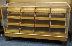 MID-CENTURY GLASS & LIGHTWOOD RETAILERS CABINET having a bank of sixteen tray-drawers, each with