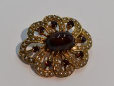A yellow metal brooch having central garnet cabouchon within an open surround of seed pearls and