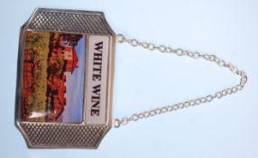 A silver plated white wine label having a square enamelled panel depicting a vineyard on a silver