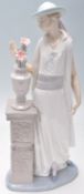 A Lladro porcelain figurine of a 1920's flapper girl modelled next to a jardiniere stand. Stamped