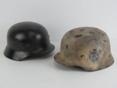 Two replica WWII helmets; white camo and black paint respectively, each with liner and chin strap.