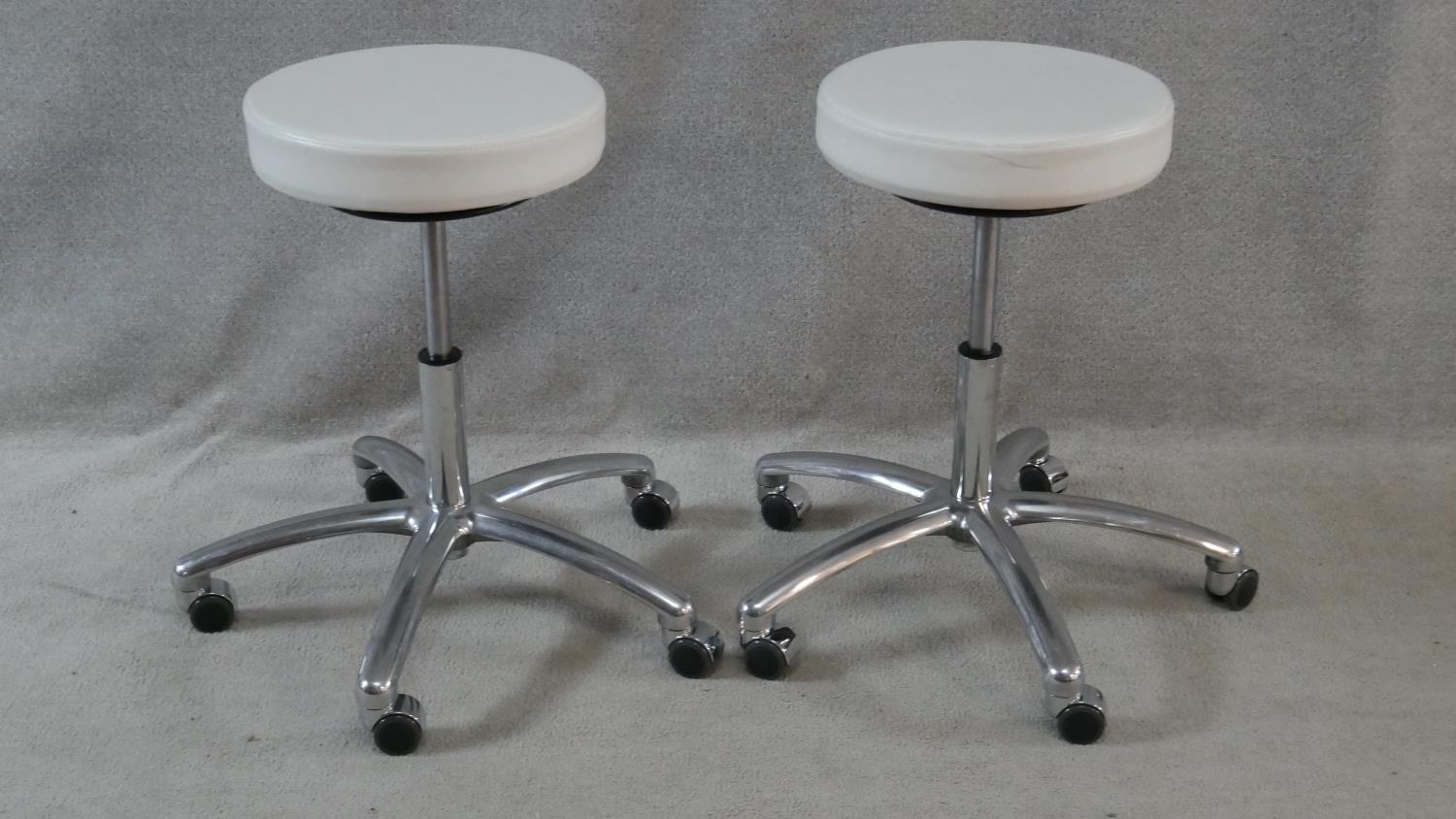 A pair of adjustable white vinyl stools on aluminium bases on casters. Makers label to the