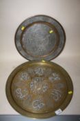 AN ISLAMIC STYLE BRASS CHARGER WITH WHITE METAL OVERLAY, TOGETHER WITH A EMBOSSED COPPER EXAMPLE,