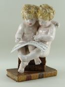 CONTINENTAL FAIENCE FIGURAL GROUP of two putti singing from a song sheet whilst seated on bookstack