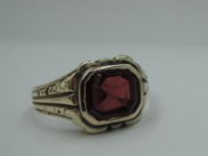 A yellow metal signet ring having a garnet style panel in collared mount to moulded shoulders on