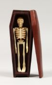 A CARVED ARTICULATED BONE SKELETON in a wooden coffin. 4.5ins long.