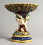 A MINTON MAJOLICA COMPORT, modelled as a pair of cherubs holding aloft a bowl. (AF) 9ins high