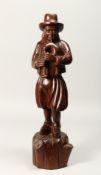 A CARVED WOOD FIGURE OF A MAN PLAYING A BLADDER PIPE. 19ins high.