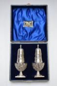 Cased Victorian pair of hallmarked silver peppers with repoussé decoration raised on square bases,