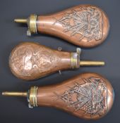 Three copper and brass powder flasks, two with embossed decoration of a cannon, guns and flags the