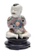 A Chinese famille verte seated figure of a smiling boy: wearing an aubergine coloured jacket
