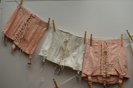 A trio of vintage boned girdles having metal suspender clasps, two pink and one white.