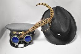 A gents vintage black trilby and steam punk cap with goggles and feathers.