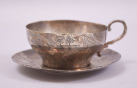 AN IRAQI WHITE METAL CUP AND SAUCER, engraved with landscape scenes.