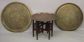 THREE LARGE ISLAMIC METAL TRAYS AND A CARVED WOOD STAND, largest tray 75cm diameter.