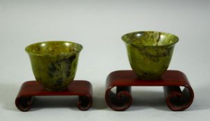 A PAIR OF CHINESE SPINACH JADE CUPS, with two wooden stands, cups 6cm diameter.