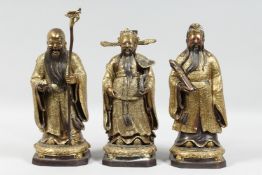THREE CHINESE GILT BRONZE IMMORTAL FIGURES, each carrying a different object, 30cm, 28.5cm and 28.