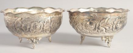 A PAIR OF WHITE METAL CIRCULAR BOWLS, with wavy rims, repousse decoration on three curving legs,