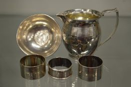 A silver milk jug, a white metal circular bowl and three silver napkin rings.