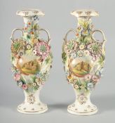 A PAIR OF FLOWER ENCRUSTED TWO HANDLED VASES painted with panels of buildings. 10.5ins high.