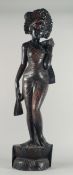 A BALINESE CARVED HARDWOOD STANDING FIGURE of a female wearing a floral headdress and stood upon a