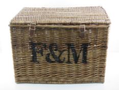 A coffee table sized wicker hamper marked 'F&M' with wicker end handles and leather straps,