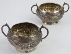 A pair of Indo-Asian white metal twin handled salts having village pastoral scenes upon with palm