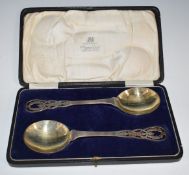 Cased pair of George V hallmarked silver serving spoons with gilt bowls and pierced finials,