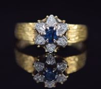 An 18ct gold ring set with a sapphire and diamonds in a flower setting, with textured shoulders, 3.