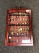 A glazed wall-hanging display cabinet containing twenty four souvenir spoons,