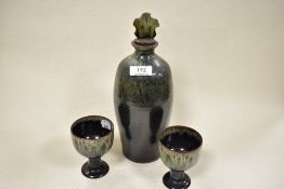 A Woburn pottery studio art decanter and two goblets, having green and brown mottled glaze.