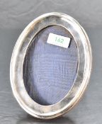 A George V silver photograph frame, of plain moulded oval form with hinged oak stand verso, marks