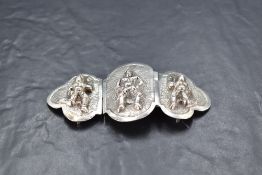 A Burmese white metal three-panel belt buckle, each panel moulded and chased with figures against