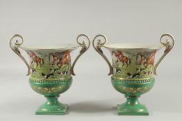 A PAIR OF SEVRES DESIGN GREEN GROUND TWO HANDLED URN SHAPED VASES with hunting scenes. 9ins high.