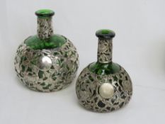 A graduated pair of emerald green glass bottles with white metal overlaid floral decoration,