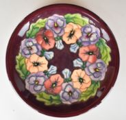 Moorcroft charger decorated in the Pansy pattern on a maroon ground, diameter 26cm