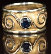 An 18ct gold bi-coloured ring set with a round cut sapphire with scrolling decoration, 5.6g, size H