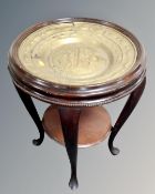 A stained beech wood two tier occasional table with brass embossed inset charger depicting Adam and