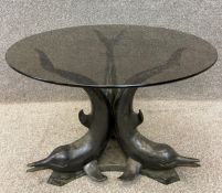 A contemporary bronze coffee table with a smoked glass circular top raised on three stylised dolphin