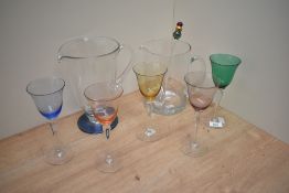 Two glass water jugs and five coloured wine glasses, in shades of green, purple, pink and yellow.