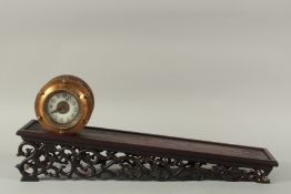 AN ORMOLU CIRCULAR INCLINE CLOCK, on a Chinese style hardwood stand. 23ins long.