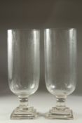 A PAIR OF GLASS STORM LANTERNS on square stepped bases. 36cms high.