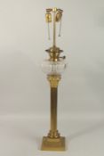 A GILT METAL CORINTHIAN COLUMN OIL LAMP with glass reservoir, on a square base. 66cms high.
