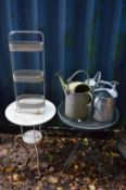 Two garden tables, three watering cans and a wrought iron plant pot stand.