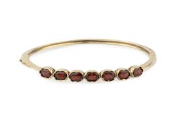A 9ct gold and garnet hinged bangle, set with seven oval cut stones and having safety chain, stamped