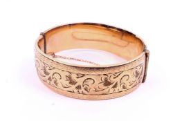 A yellow metal hinged bangle. Having engraved decoration and a clip-lock clasp with a safety chain.
