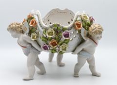 A large porcelain group of winged Cherubs carrying a basket incrusted with flowers with cross swords