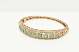 A GILT HINGED BANGLE, designed with a row of eleven oval cut emeralds, each in a four claw