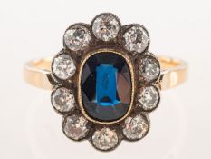 A sapphire and diamond cluster ring, collet and milgrain set with an oval-cut dark blue sapphire,